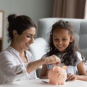 A woman puts coins into a piggy bank next to a child.